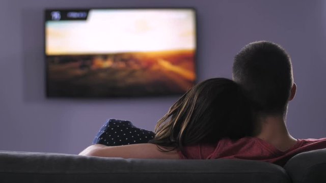 Back View Of Young Adults Couple Sitting On The Sofa Together And Watching Plazma TV At Night. Embracing Boyfriend And Girlfriend Relaxing Peacefully Together While Man Changing Tv Channels.