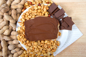 Chocolate peanutbutter spread on bread, open face sandwich on plate with pieces of chocolate, shelled peanuts and whole in shell peanuts on wood table. Tasty combination of chocolate and peanuts