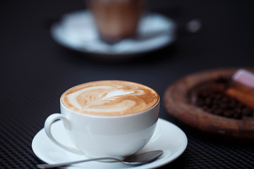 Hot cappuccino art with white cup on coffee house. Part of glass beaker and round wooden plate on...