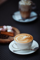 Hot Cappuccino art with white cup on coffee house. Blurry boozy cocktail with whipped cream and round wooden plate on dark background. Copy space