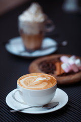 Hot Cappuccino art with white cup on coffee house. Blurry boozy cocktail with whipped cream and round wooden plate on dark background. Vertical, copy space
