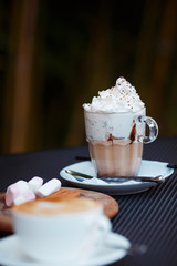 Cozy mug with cream. Boozy cocktail with whipped cream on top and chocolate chips. Round wooden plate with cinnamon roll, coffee beans and sugar pads. Blurry cappuccino on the foreground.
