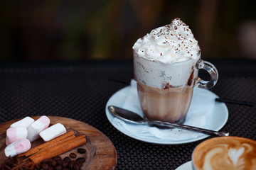 Cup of cozy mug with cream. Boozy cocktail with whipped cream on top and chocolate chips. Round wooden plate with cinnamon roll, coffee beans and sugar pads. Blurry cappuccino on the foreground