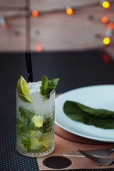 Mojito cocktail with lime and mint in highball glass on a grey table. Blurred LED lights and round plate with greeen leaf on background. Copy Space