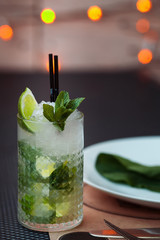 Mojito cocktail with lime and mint in highball glass on a grey table. Blurred LED lights and round plate with greeen leaf on background. Copy Space