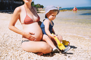 Pregnant mother and toddler child relaxing on beach at summer