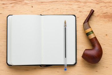 Ballpoint pen on a notebook and a pipe next to it on a wooden table