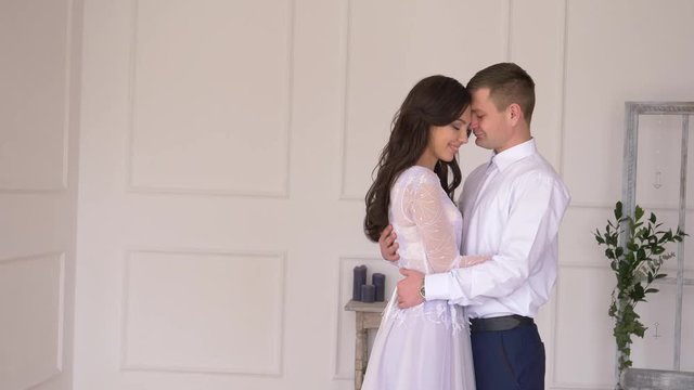young couple against white wall background, man in white shirt girl with braces in tender dress serene look at each other and hug.