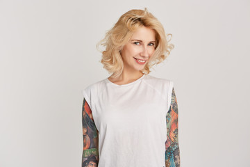 Shy and smiling blonde girl with earring in nose and tattoes over her arms looking directly in camera. Isolated over white wall