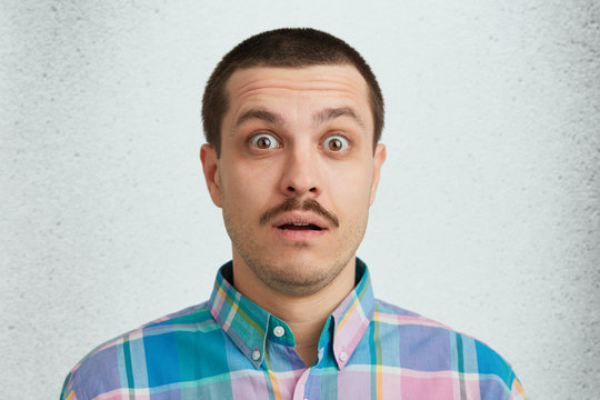 Portrait of unshaved surprised astonished hesitant male with dark warm eyes and mustaches wearing colorful shirt being shoked with last news. Isolated over studio white wall