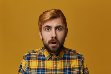 Portrait of stupefied bearded male, with dyed blond gold hair keeps mouth widely opened, wonders about something, isolated over yellow background