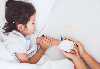 Mother hand holding sick daughter hand who have IV solution bandaged with love and care while she is sleeping on bed in the hospital