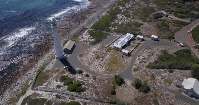 4K high quality aerial video sunny day view of historical Slangkop lighthouse built in 1914 and Kommetjie town in the background near beach at sea shore in Western Cape near Cape Town, South Africa