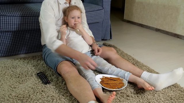 man and daughter watching television, sitting on the floor eating snacks