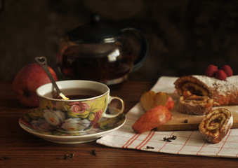 A cup of tea, a glass teapot, a sweet roll and pieces of apples on a wooden table. Still life.