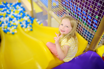 Fototapeta na wymiar Happy little girl having fun in ball pit in kids indoor play center. Child playing with colorful balls in playground ball pool.