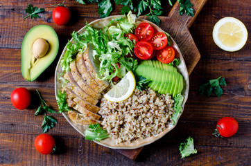 Healthy salad bowl with quinoa, tomatoes, chicken, avocado, lime and mixed greens, lettuce, parsley on wooden background close up. Food and health.