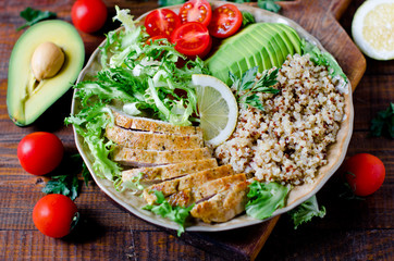 Healthy salad bowl with quinoa, tomatoes, chicken, avocado, lime and mixed greens, lettuce, parsley on wooden background close up. Food and health.