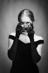 Beautiful girl in evening dress and long gloves with a camera. Black and white portrait in studio