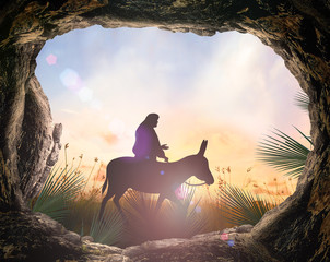 Easter Religious concept: Silhouette Jesus Christ riding donkey with tomb stone on meadow sunset background