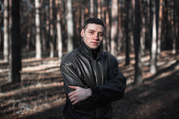 A young man of criminal appearance in a black leather jacket posing in an autumn forest.