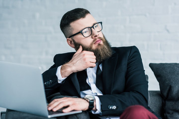 portrait of pensive businessman in eyeglasses with laptop looking away