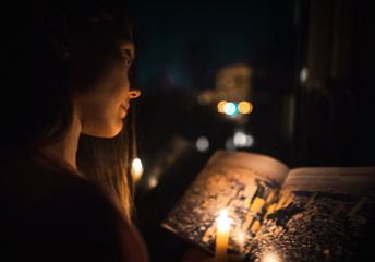 A beautiful young girl in a shirt reading a book at the window-sill by the light of a burning candle at night