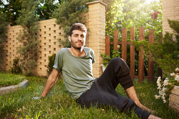 Outdoors portrait of young attractive bearded caucasian male gardener smiling, sitting on grass in...