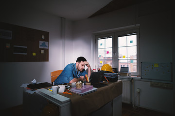 Young exhausted disappointed business man searching for problem solution leaning his head on hand while staying after regular working time in the dark office.