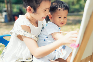 Fototapeta premium Asian little boy and sister draw together in park. Kids painting and drawing. Children paint with paintbrush color and pencils. Art and crafts for toddler and preschooler..