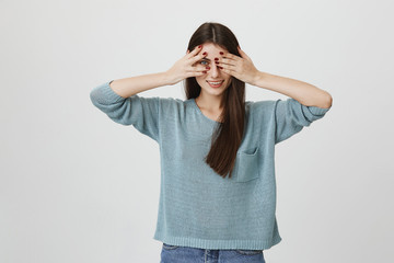 Indoor shot of young beautiful caucasian woman covering her eyes with hands while smiling and peeking to see something in front, standing over gray background. Girl is excited and can not wait to look