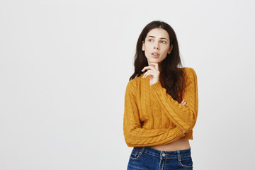 Portrait of intrigued charming european girl, standing in thoughtful pose with opened mouth, looking aside and holding hand on ching, planning or calculating something in mind, over gray background.