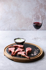 Grilled sliced rack of lamb with yogurt mint sauce served with green salad young beetroot leaves, glass of red wine, pink salt on round wooden slate board over grey texture table. Copy space