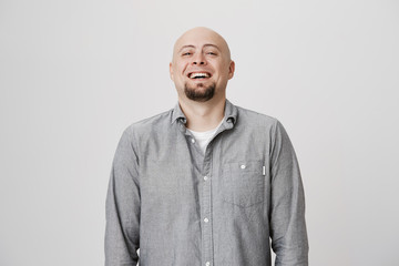 Portrait of attractive bald man laughing and smiling standing near white wall. Positive emotions concept. Guy feels happy and enjoys spending time in such funny and pleasant company.