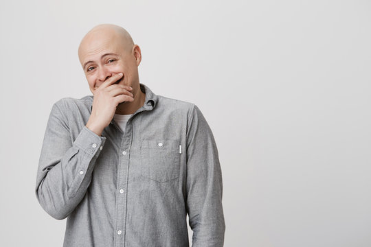 Close up portrait of bald man laughing and covering his mouth with hand over white background. Attractive guy finds something really funny but he tries to hold his laugh not to offend some person.