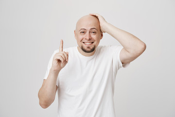 Studio portrait of attractive single bald man with beard touching head while pointing up and smiling while standing against gray background. Man finally remembered or got interesting idea