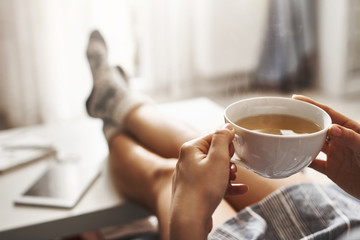 Cup of tea and chill. Woman lying on couch, holding legs on coffee table, drinking hot coffee and enjoying morning, being in dreamy and relaxed mood. Girl in oversized shirt takes break at home - 193565473