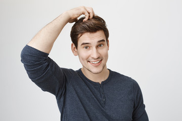 Portrait of funny handsome caucasian man in casual clothes acting like monkey, smiling and scratching head, standing against gray background. Boy attend acting classes in local theatre