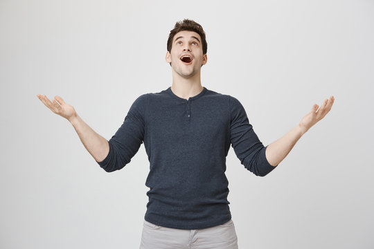 Portrait of fascinated excited young guy looking up with happy expression and raising arms, standing over gray background. Boyfriend sings to gilfriend while standing near her balcony
