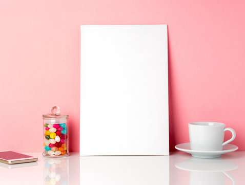 Blank white frame and candys in jar, cup of coffee or tea on a white table against the pink wall with copy space
