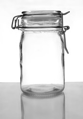 A hermetic sealing jar that is empty sitting on a reflective grey surface with bright background. The edges are enhanced by using a technique called bright field lighting. The jar is closed.
