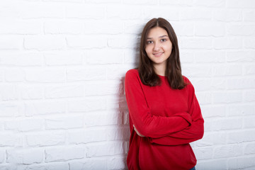 A young pretty smiling girl in a red sweater and a hat against a modern white brick wall in the loft style. She looks at the viewer. Copy space.
