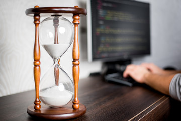 Hourglass or Sandglass at a desk showing time flow while a guy working on computer on a background. Time concept. Focus on a sand with a copy space