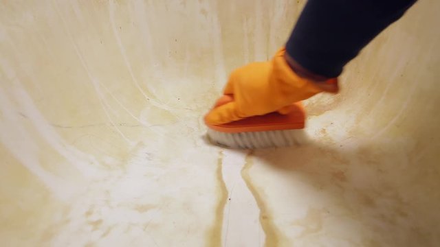 A man's hand in orange rubber gloves cleaning the bathtub sink with a scrub brush. General house cleaning.