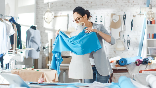 Beautiful Female fashion, designer,  Adjusting Fabrics on a Tailored Mannequin. In Her Studio Various Sewing Items and Colorful Fabrics Laying around, Mannequins Standing, and Sketches Pinned.