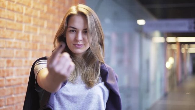 Beautiful young woman looking at camera, smiling and pointing with her finger. She is calling the viewer while standing in a building corridor with brick walls. Handheld slow motion medium shot