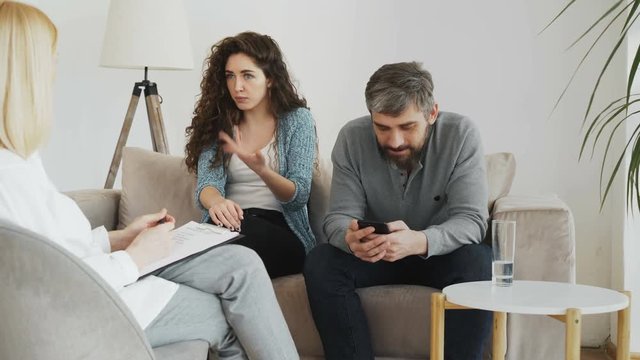 Female professional psychologist listening and writing notes while young woman arguing and her boyfriend surfing in smartphone during visit psychotherapy office indoors