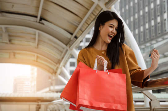 beautiful cheerful asian holding many colorful red shopping bags and credit card walking in modern big city, fashion, promotion, special offer, winter sale, people, payment and shopping online concept