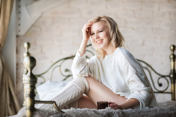 Obraz premium Beautiful photo of a sexy woman in the morning. Young girl in the morning in bed in a white sweater and with a cup of tea in hands. Blonde girl with short hair smiles.