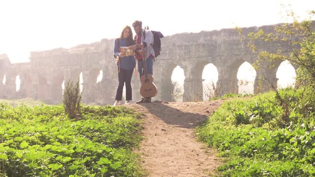 Young lovely couple backpackers tourists reading map pointing directions roman aqueduct arches in parco degli acquedotti park ruins in rome on romantic misty sunrise with guitar and sleeping bag slow